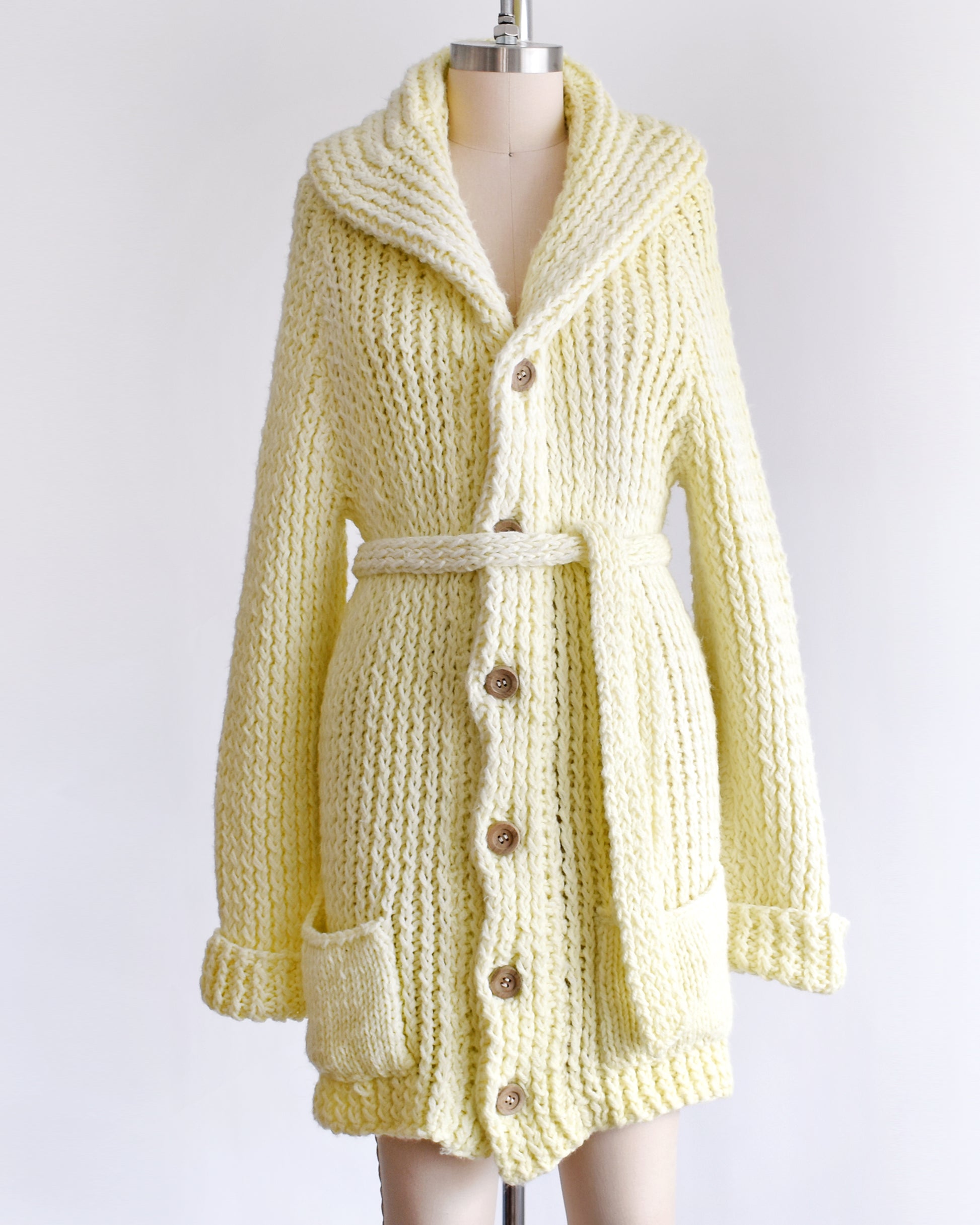 A vintage 1970s cream chunky knit cardigan with wood buttons down the front with a matching tie belt on a dress form.