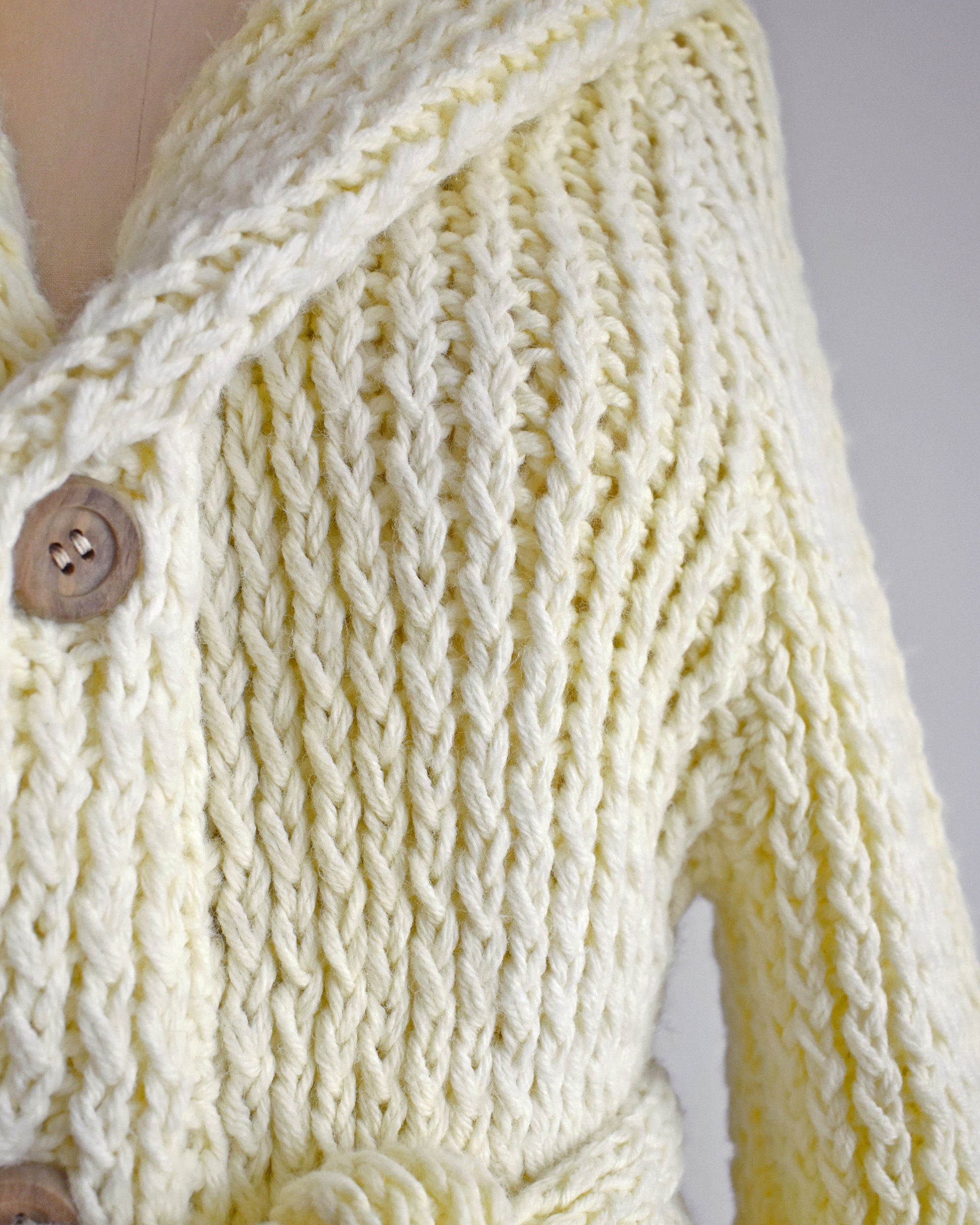 close up of the bodice of a a vintage 1970s cream chunky knit cardigan with wood buttons down the front with a matching tie belt on a dress form. Some spots of the knit are lighter than the rest.