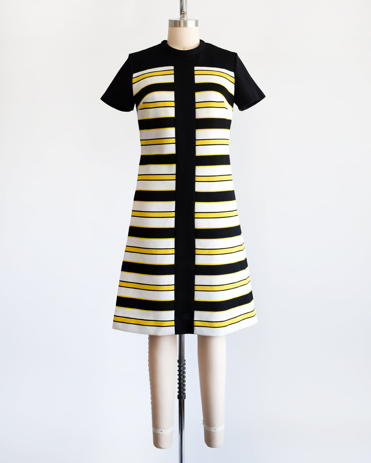 A vintage 1960s short-sleeve knit dress displayed on a mannequin against a plain white background. The dress features a black yoke and sleeves, with horizontal black, white, and yellow stripes 