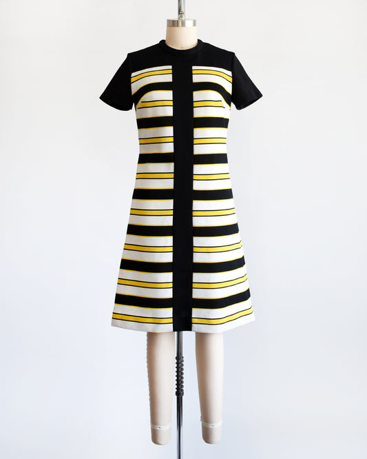 A vintage 1960s short-sleeve knit dress displayed on a mannequin against a plain white background. The dress features a black yoke and sleeves, with horizontal black, white, and yellow stripes 
