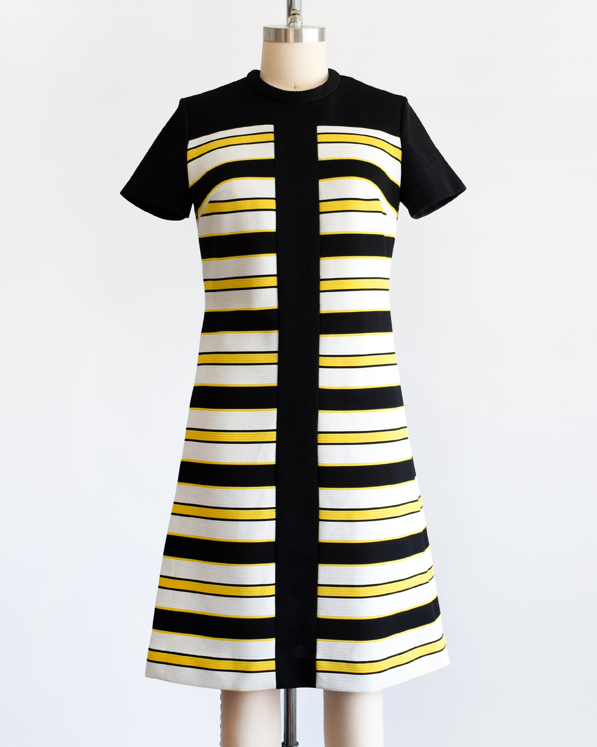 A vintage 1960s short-sleeve knit dress displayed on a mannequin against a plain white background. The dress features a black yoke and sleeves, with horizontal black, white, and yellow stripes 
