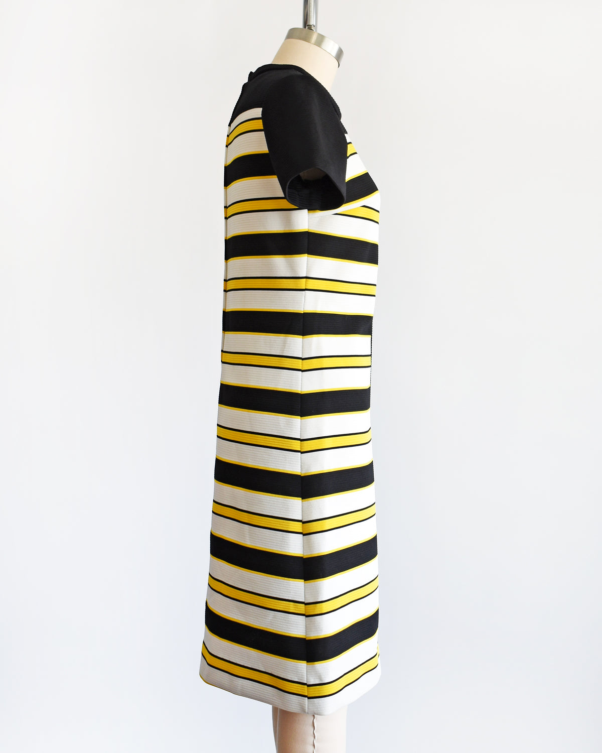 side view of a vintage 1960s short-sleeve knit dress displayed on a mannequin against a plain white background. The dress features a black yoke and sleeves, with horizontal black, white, and yellow stripes 