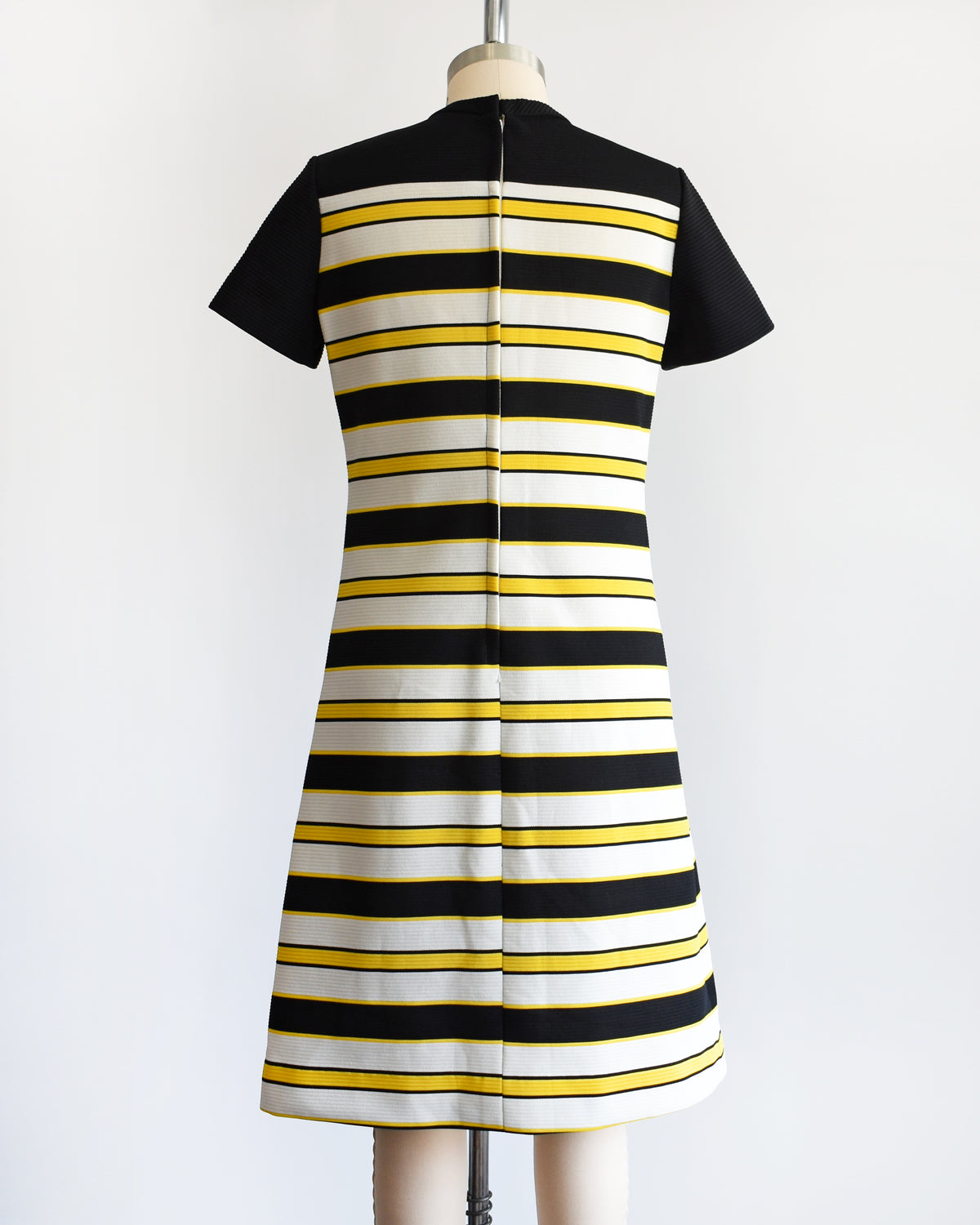 back view of a vintage 1960s short-sleeve knit dress displayed on a mannequin against a plain white background. The dress features a black yoke and sleeves, with horizontal black, white, and yellow stripes 
