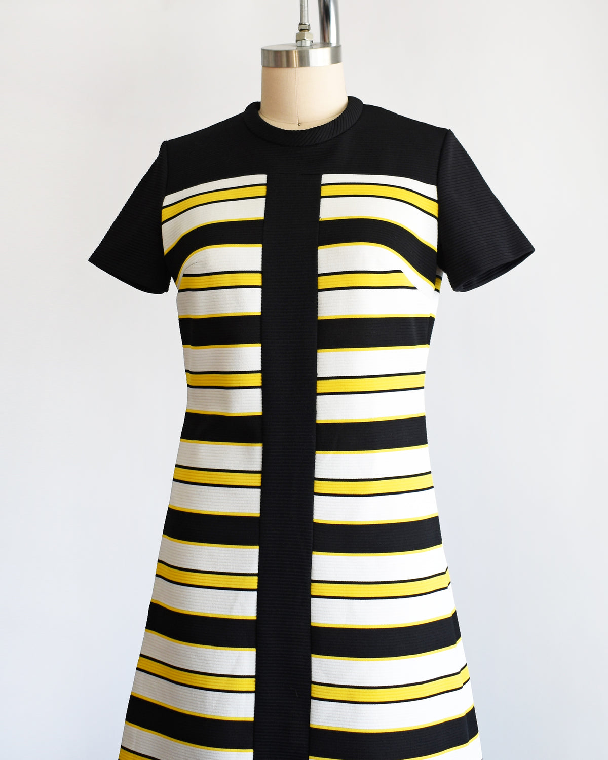 side front view of a vintage 1960s short-sleeve knit dress displayed on a mannequin against a plain white background. The dress features a black yoke and sleeves, with horizontal black, white, and yellow stripes 