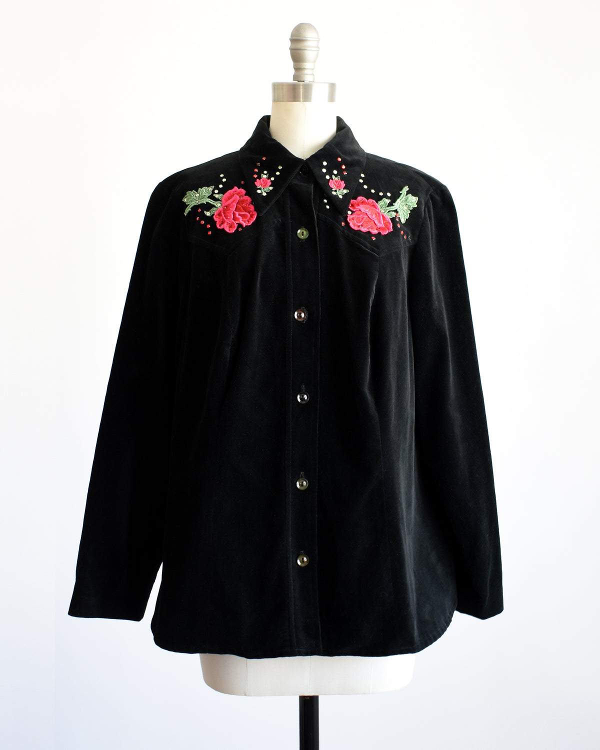 A vintage black long-sleeve button-front shirt jacket displayed on a dress form mannequin. The jacket features embroidered red roses with green leaves and small rhinestone accents on the chest.