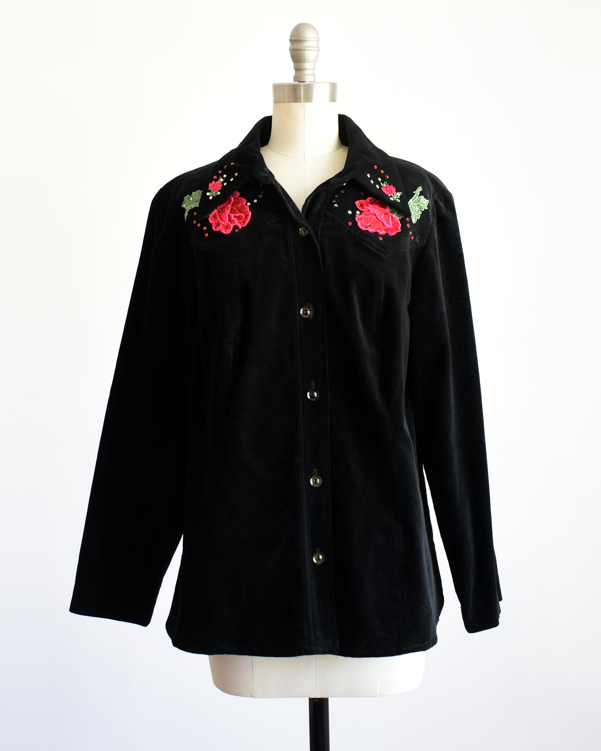 A vintage black long-sleeve button-front shirt jacket displayed on a dress form mannequin. The jacket features embroidered red roses with green leaves and small rhinestone accents on the chest.