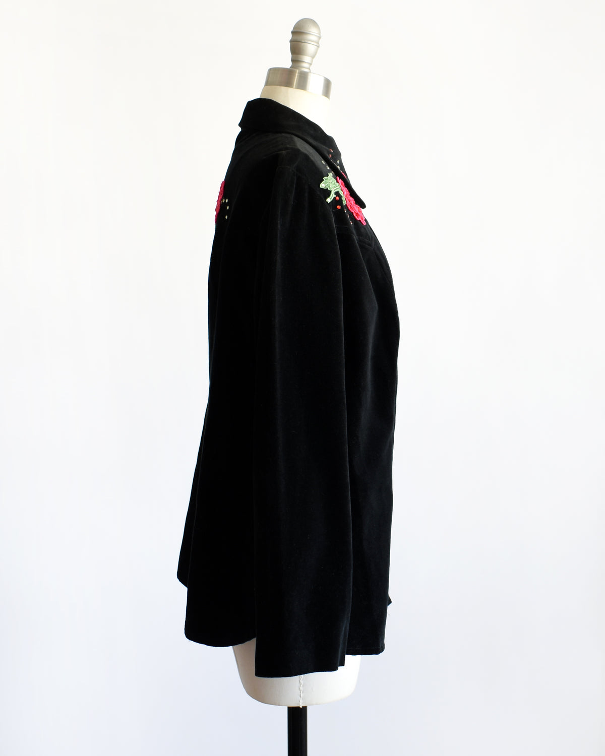 side view of a vintage black long-sleeve button-front shirt jacket displayed on a dress form mannequin. The jacket features embroidered red roses with green leaves and small rhinestone accents on the chest.