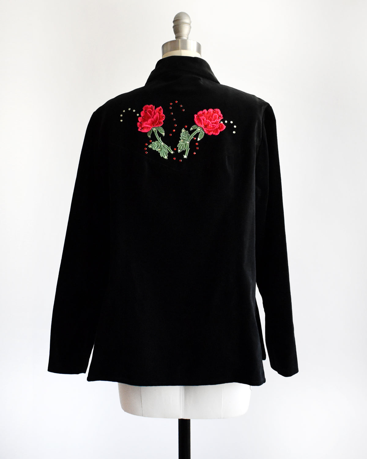 back view of a vintage black long-sleeve button-front shirt jacket displayed on a dress form mannequin. The jacket features embroidered red roses with green leaves and small rhinestone accents on the chest.