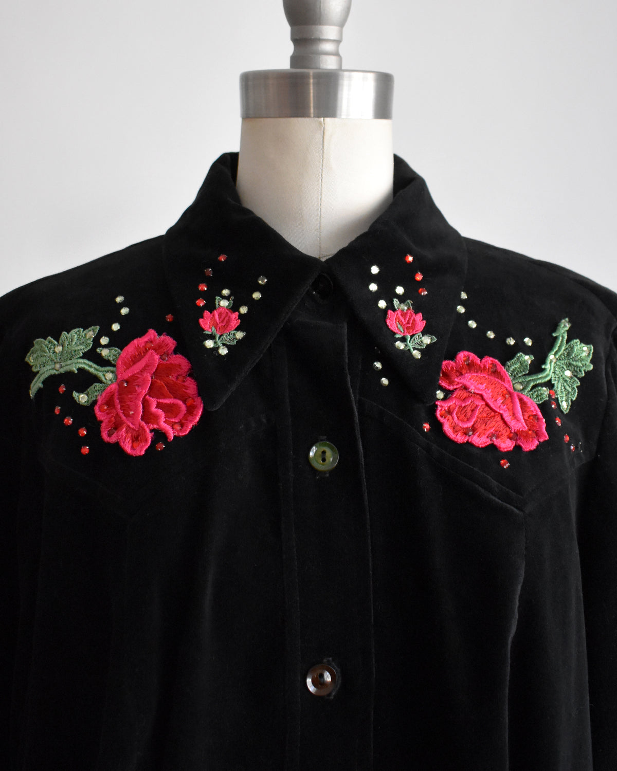 close up of the collar on a vintage black long-sleeve button-front shirt jacket displayed on a dress form mannequin. The jacket features embroidered red roses with green leaves and small rhinestone accents on the chest.