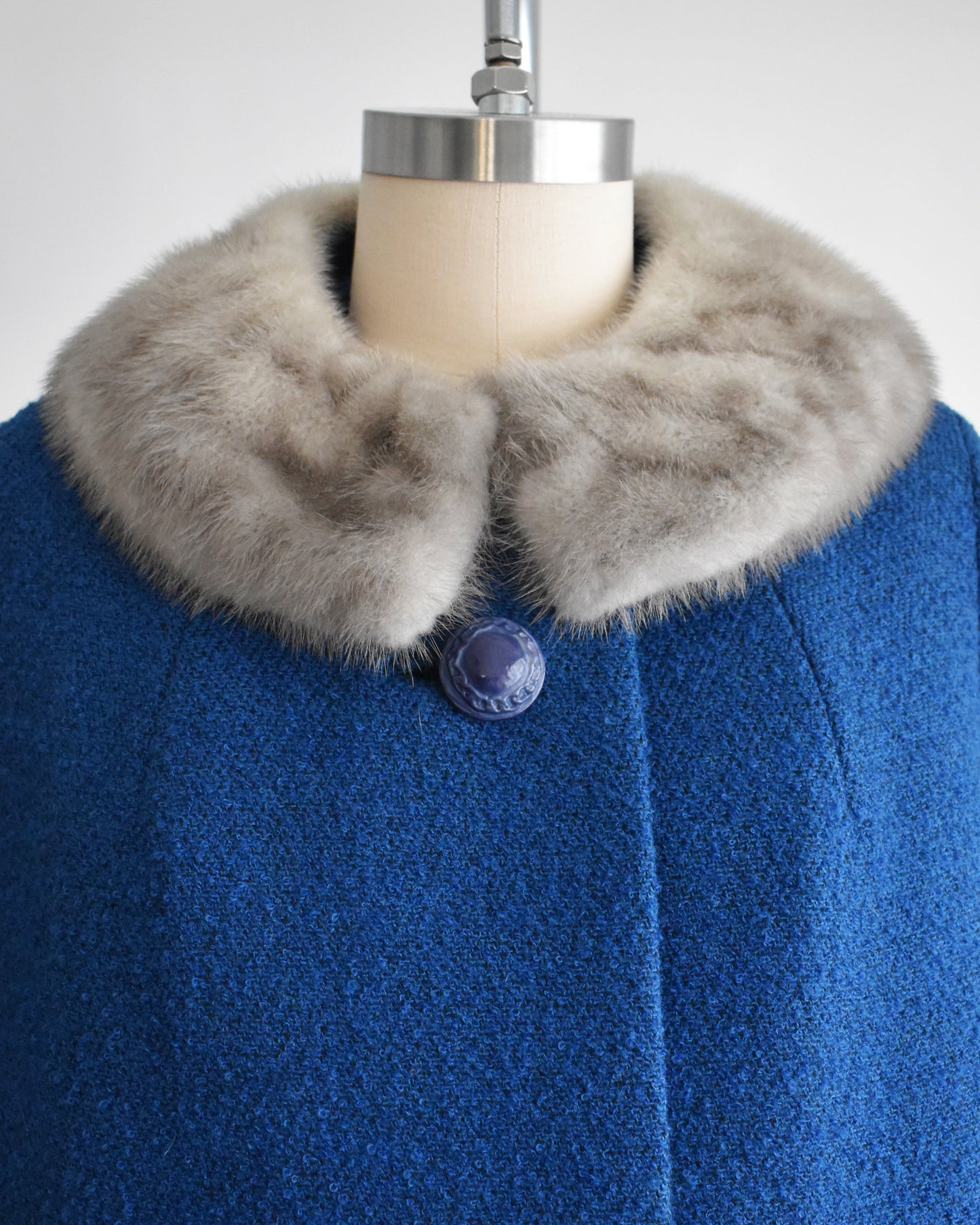 close up of the fur collar and top button on a vintage 1960s blue wool coat with gray fur trimmed collar on a mannequin against a white background