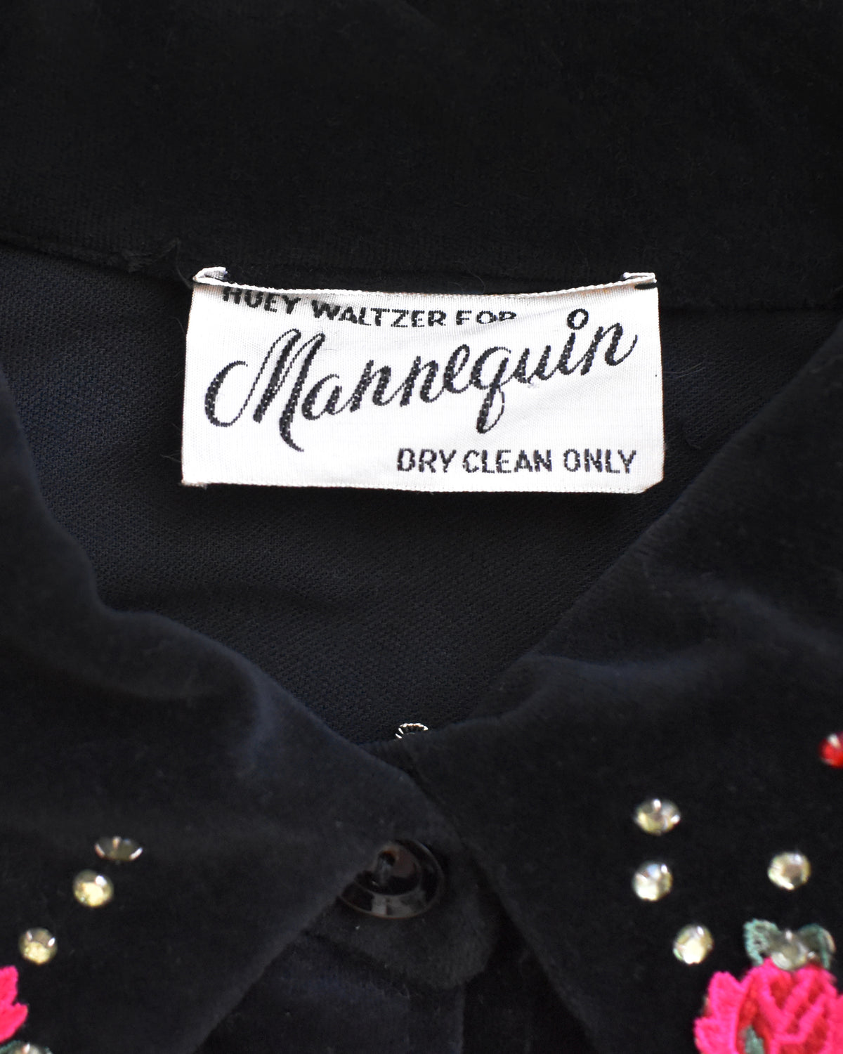 Close-up of a black velvet garment label reading 'Huey Waltzer For Mannequin Dry Clean Only' on a black fabric surface with small floral and rhinestone embellishments visible.