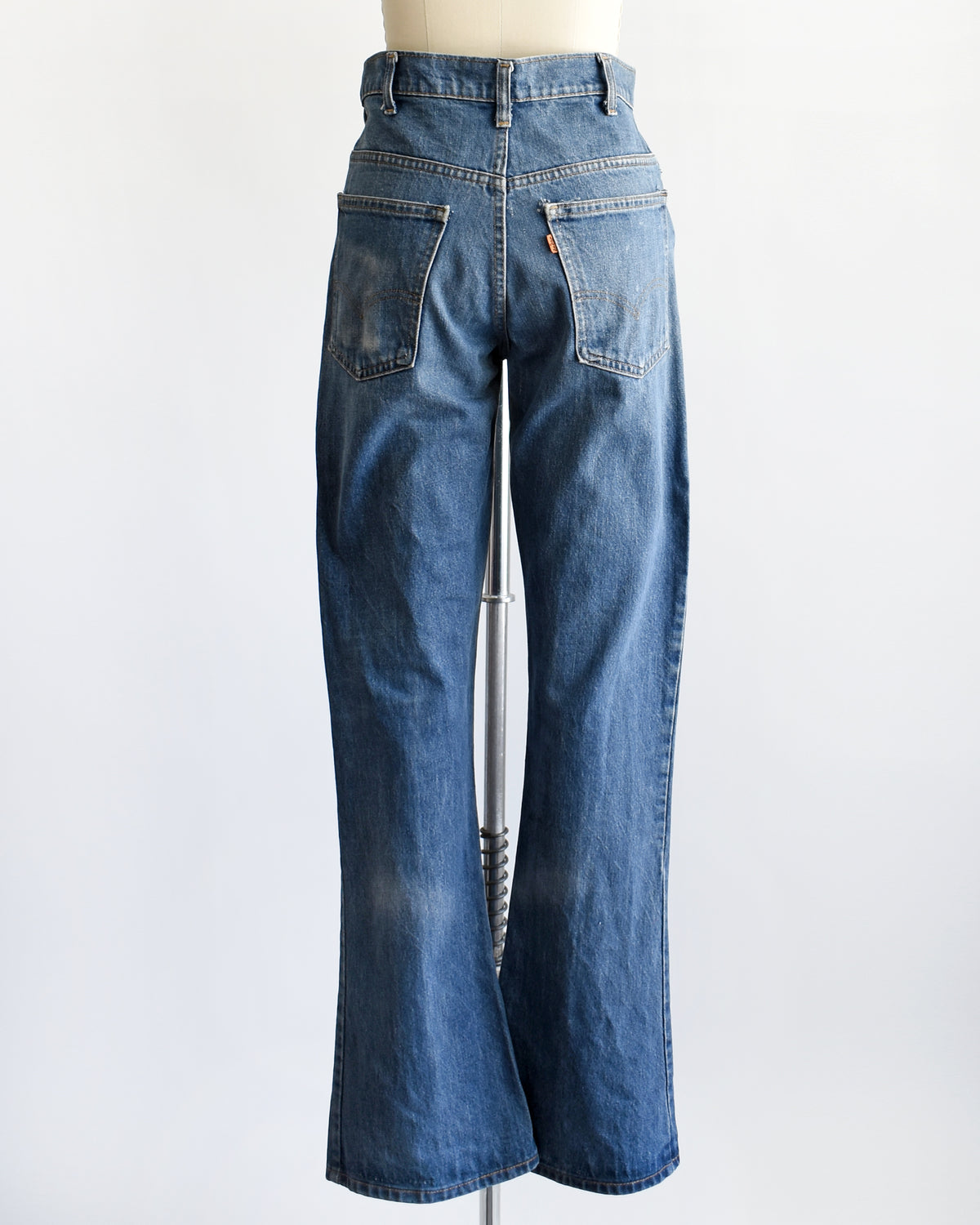 back view of a pair of medium-wash blue denim flare vintage Levi's jeans displayed on a cream-colored dress form mannequin against a plain white background.