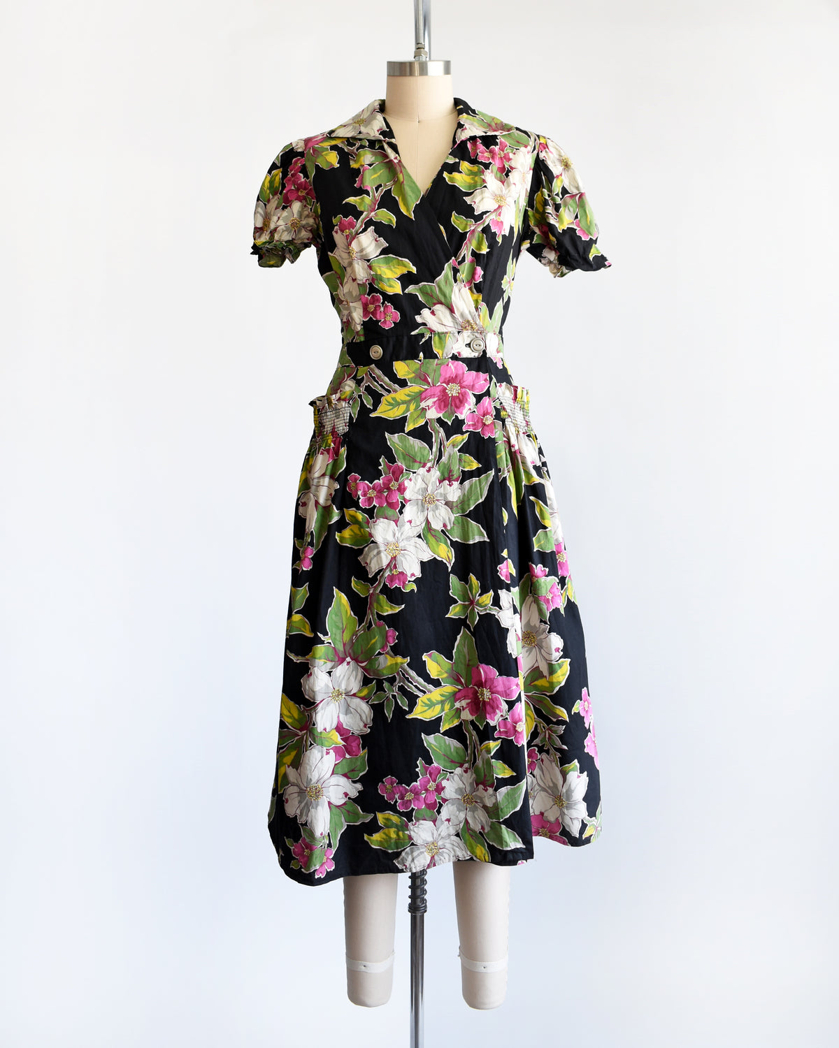 A vintage 1940s/1950s black midi dress with a vibrant pink, white, and green floral print, displayed on a cream mannequin against a plain white background.