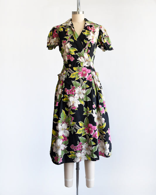 A vintage 1940s/1950s black midi dress with a vibrant pink, white, and green floral print, displayed on a cream mannequin against a plain white background.