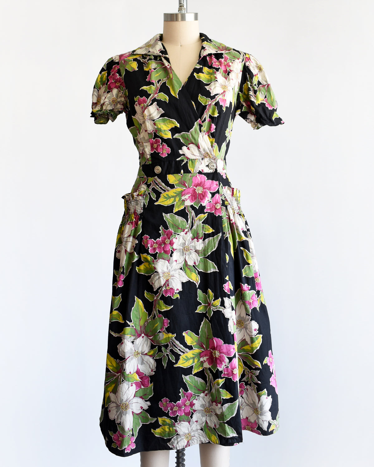 A vintage 1940s/1950s black midi dress with a vibrant pink, white, and green floral print, displayed on a cream mannequin against a plain white background.