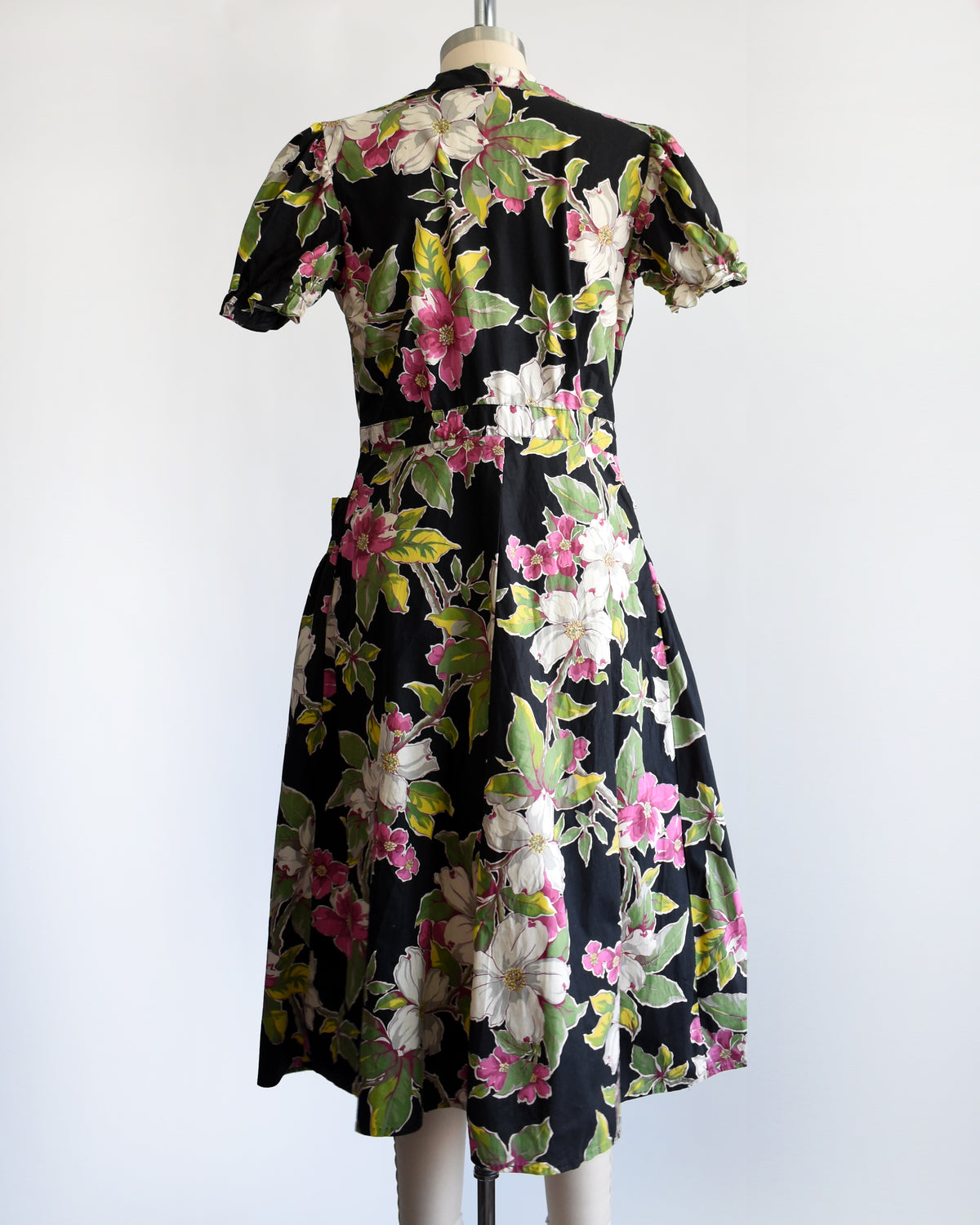 back view of a vintage 1940s/1950s black midi dress with a vibrant pink, white, and green floral print, displayed on a cream mannequin against a plain white background.