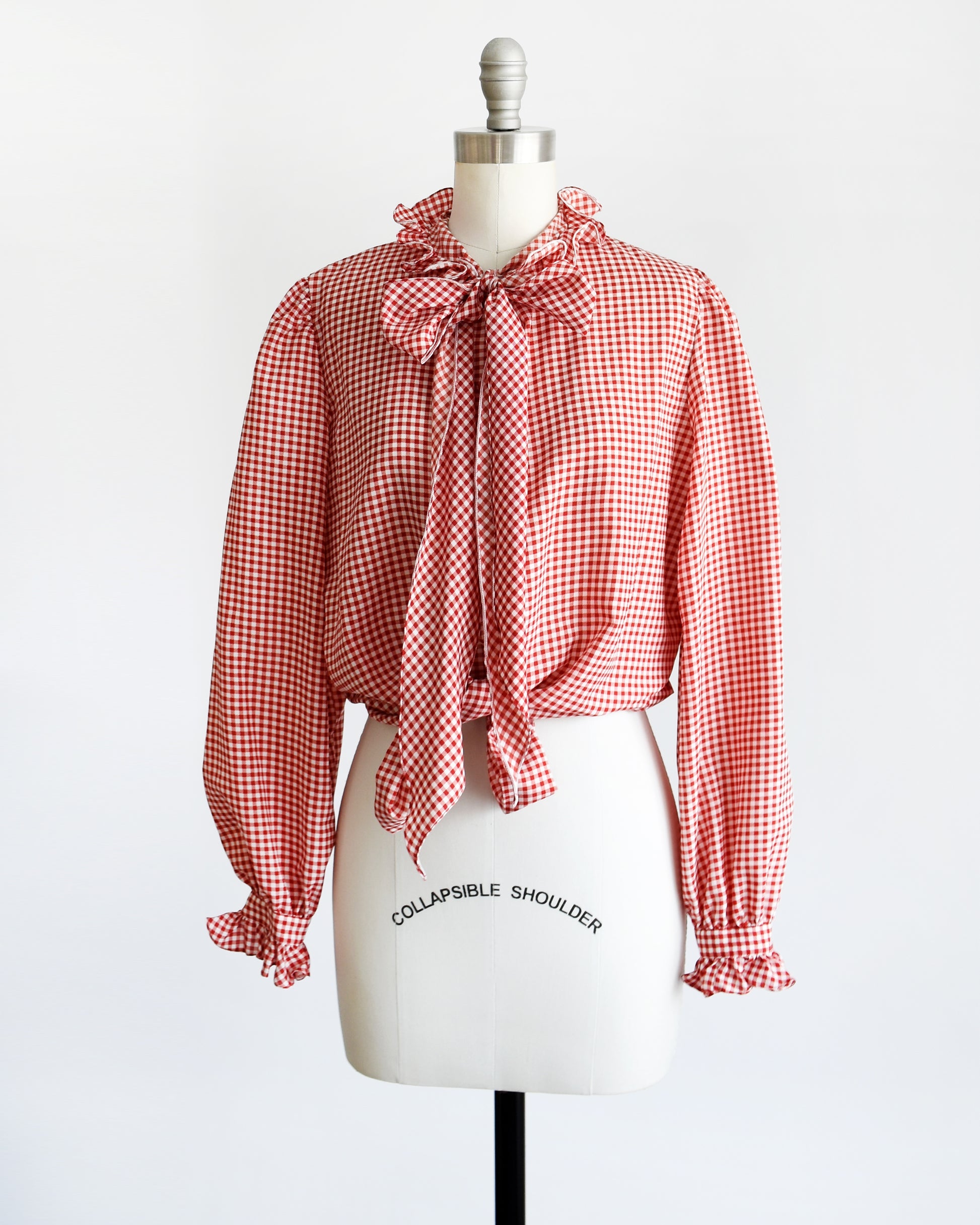 Side front view of a vintage 1980s red and white gingham blouse with ruffled collar and ascot bow. The blouse is tied at the bottom giving it a cropped look