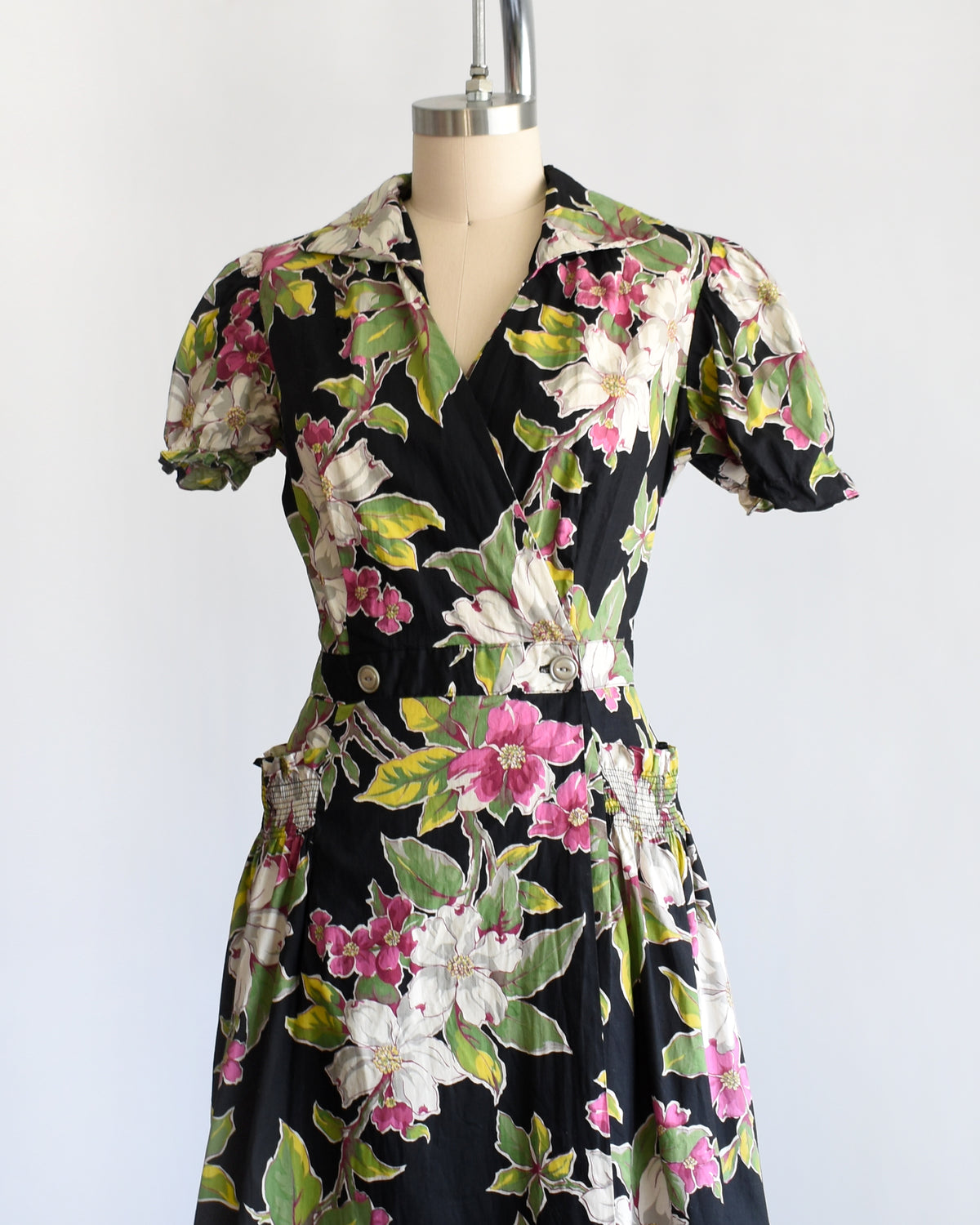 side front view of a vintage 1940s/1950s black midi dress with a vibrant pink, white, and green floral print, displayed on a cream mannequin against a plain white background.