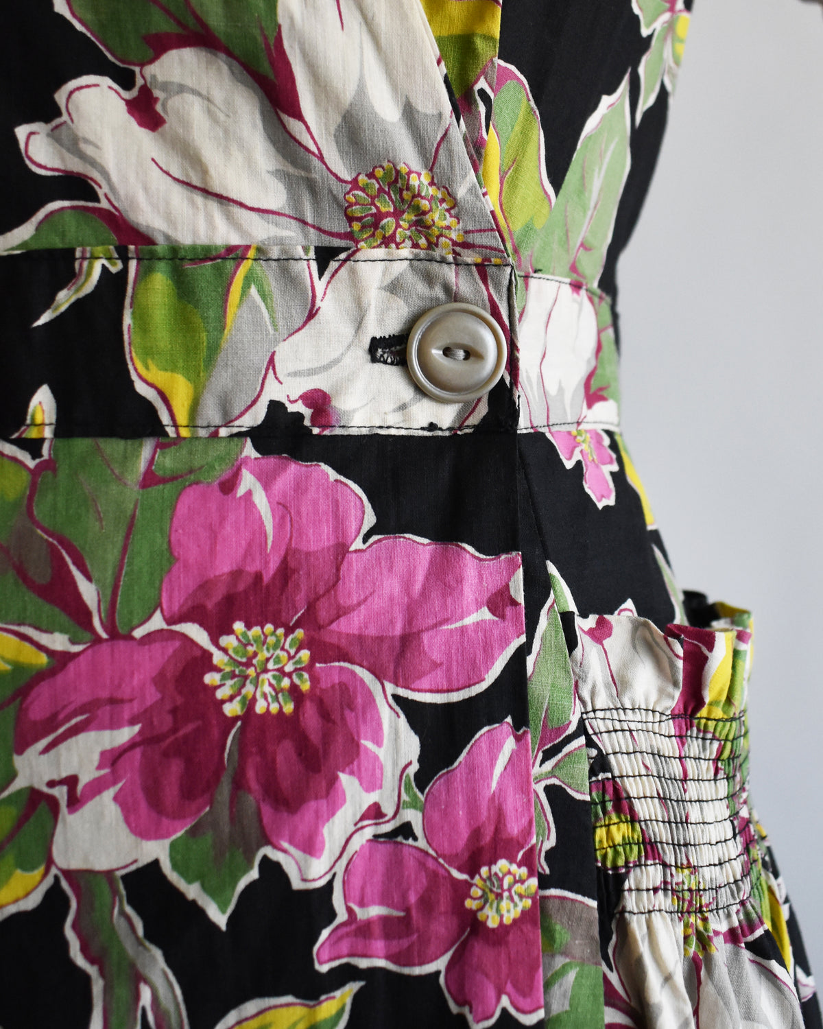 Close-up of the waist of the dress which features a large pink and white flowers with green leaves on a black background, with a visible button and smocked fabric detail.