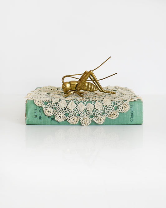 A vintage brass grasshopper/cricket on a lace doily on a green book.