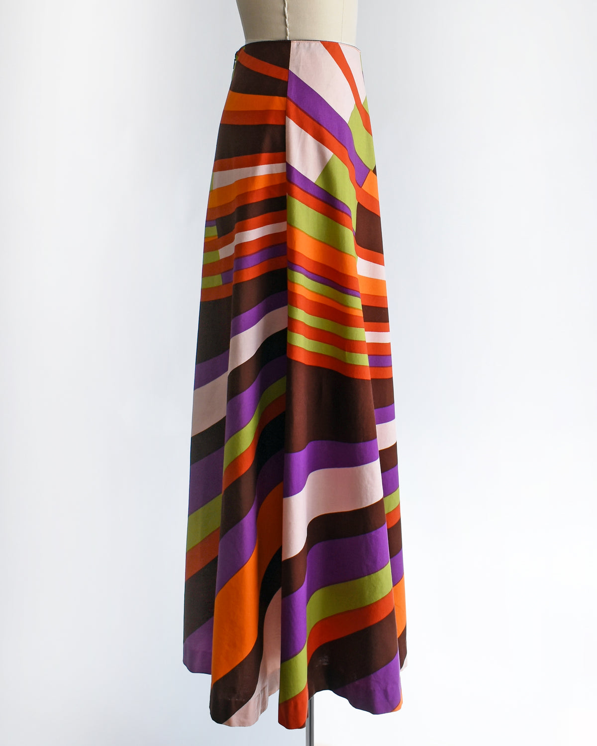 side view of a high-waisted vintage 1974 Marimekko A-line midi skirt with a bold retro diagonal stripe pattern in orange, purple, brown, green, and cream, displayed on a mannequin against a white background.
