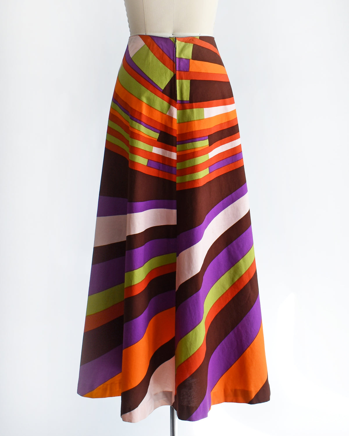 back view of a high-waisted, vintage 1974 Marimekko A-line midi skirt with a bold retro diagonal stripe pattern in orange, purple, brown, green, and cream, displayed on a mannequin against a white background.