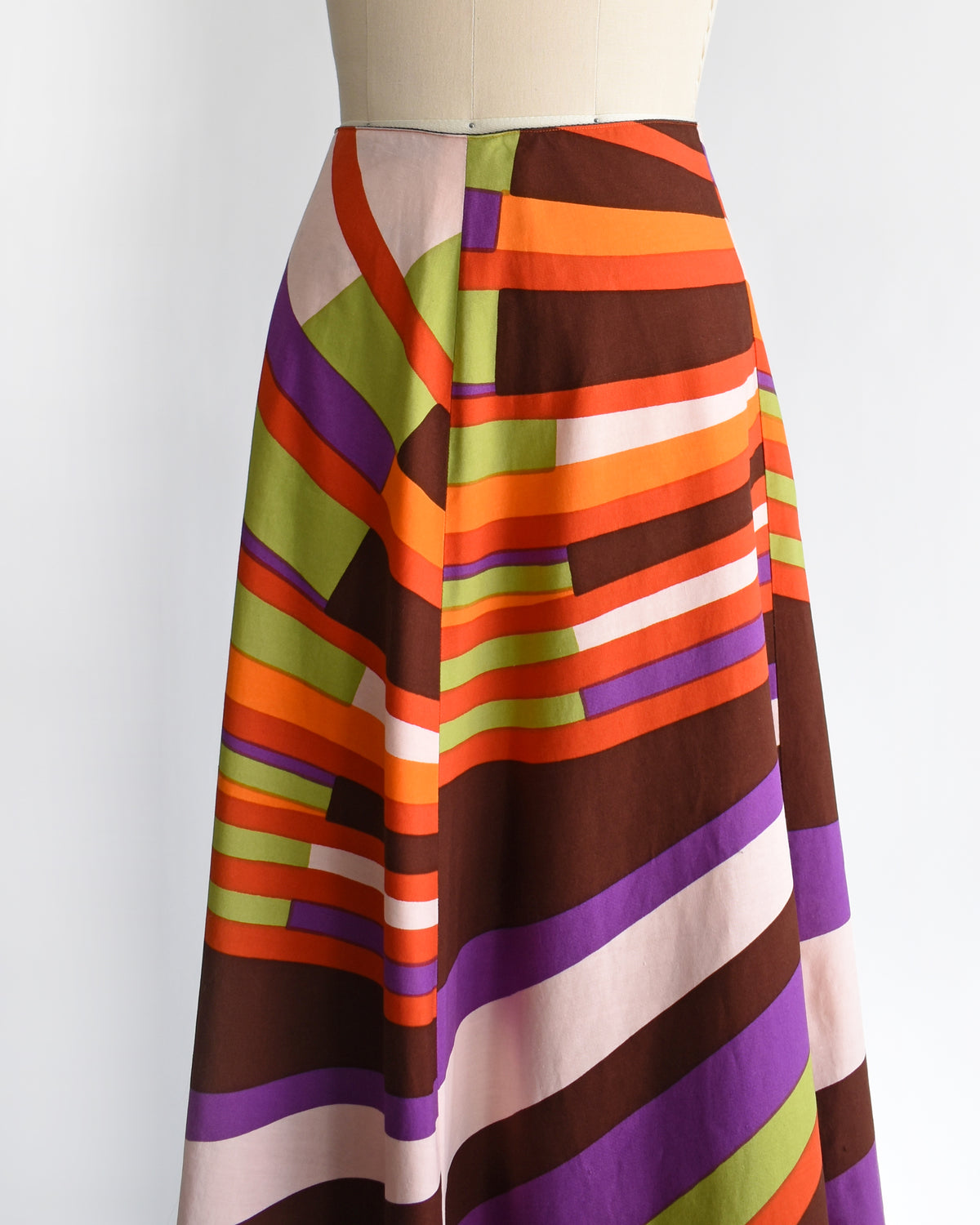 side front view of a high-waisted, vintage 1974s Marimekko A-line midi skirt with a bold retro diagonal stripe pattern in orange, purple, brown, green, and cream, displayed on a mannequin against a white background.