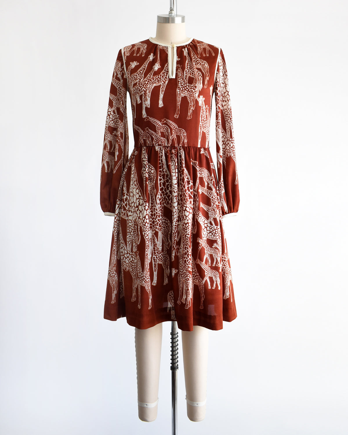 A vintage 1970s long-sleeve A-line dress displayed on a mannequin against a plain white background. The dress features a rust-brown base with a white giraffe print and a keyhole neckline with button details.