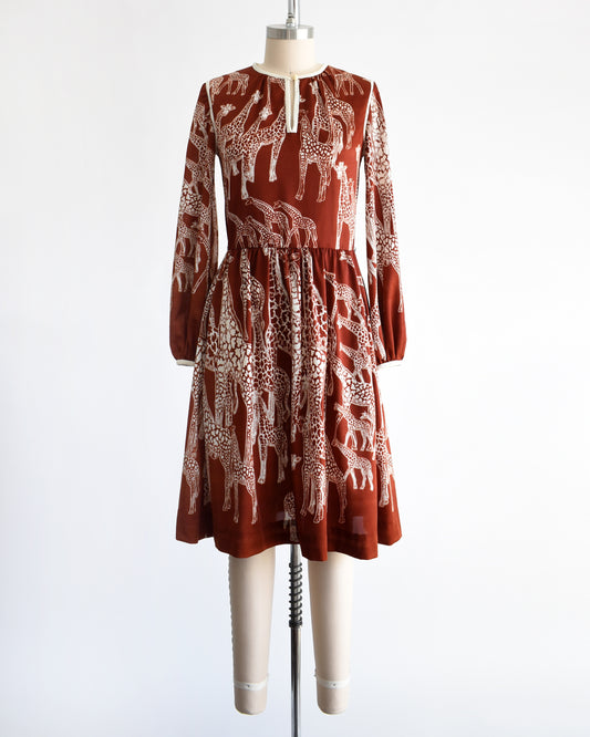 A vintage 1970s long-sleeve A-line dress displayed on a mannequin against a plain white background. The dress features a rust-brown base with a white giraffe print and a keyhole neckline with button details.