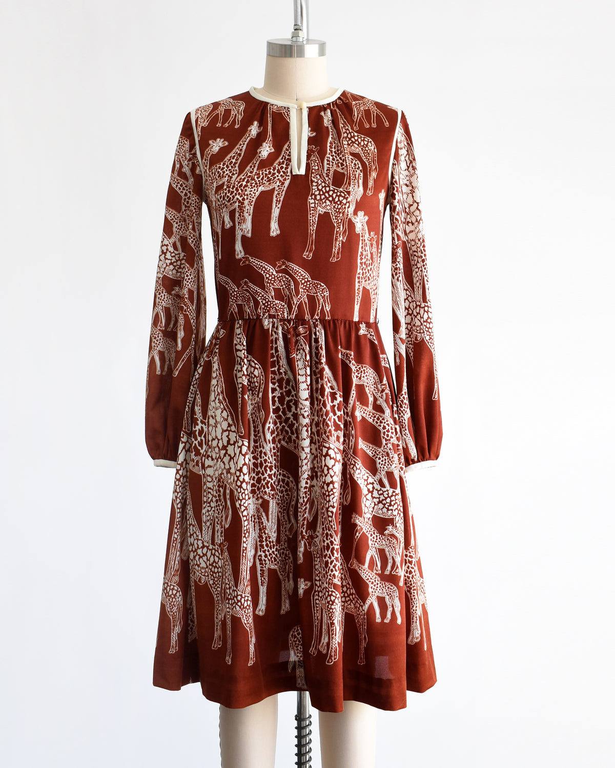 A vintage 1970s long-sleeve A-line dress displayed on a mannequin against a plain white background. The dress features a rust-brown base with a white giraffe print and a keyhole neckline with button details.
