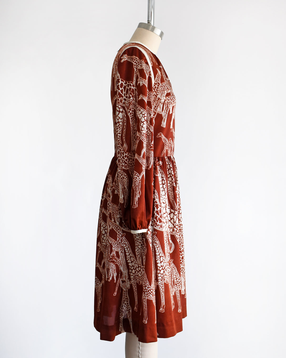 side view of a vintage 1970s long-sleeve A-line dress displayed on a mannequin against a plain white background. The dress features a rust-brown base with a white giraffe print and a keyhole neckline with button details.