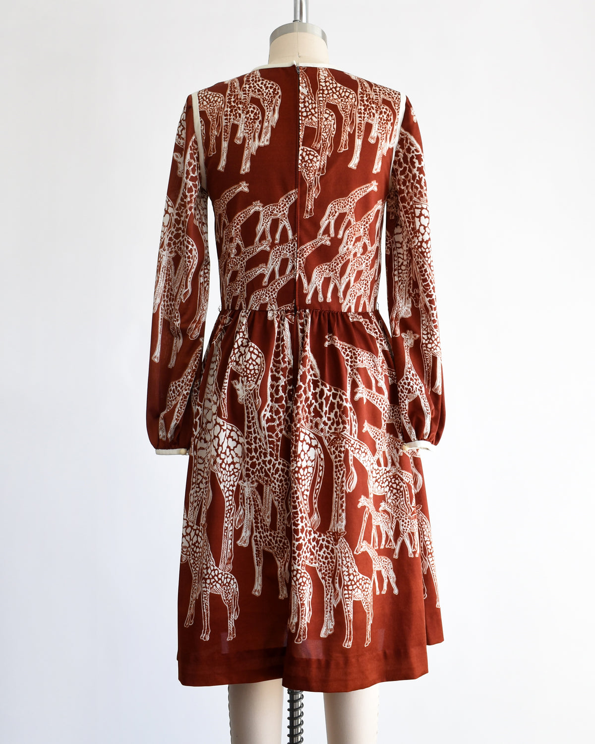 back view of a vintage 1970s long-sleeve A-line dress displayed on a mannequin against a plain white background. The dress features a rust-brown base with a white giraffe print and a keyhole neckline with button details.