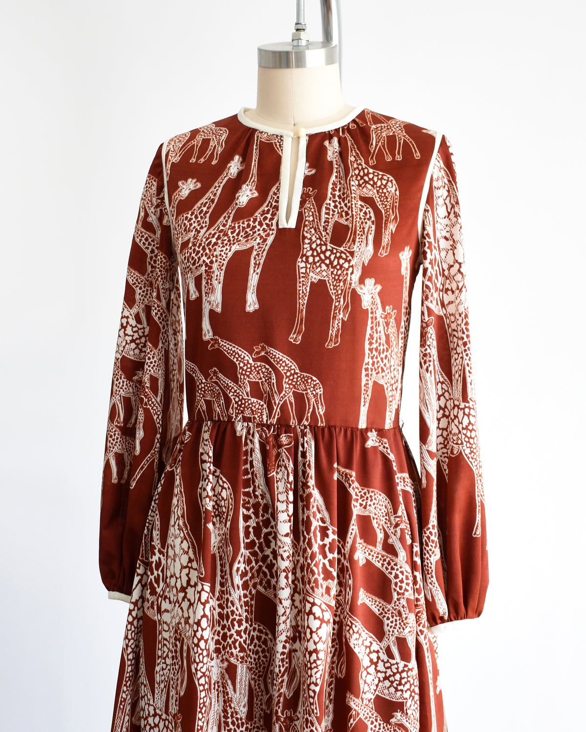 side front view of a vintage 1970s long-sleeve A-line dress displayed on a mannequin against a plain white background. The dress features a rust-brown base with a white giraffe print and a keyhole neckline with button details.