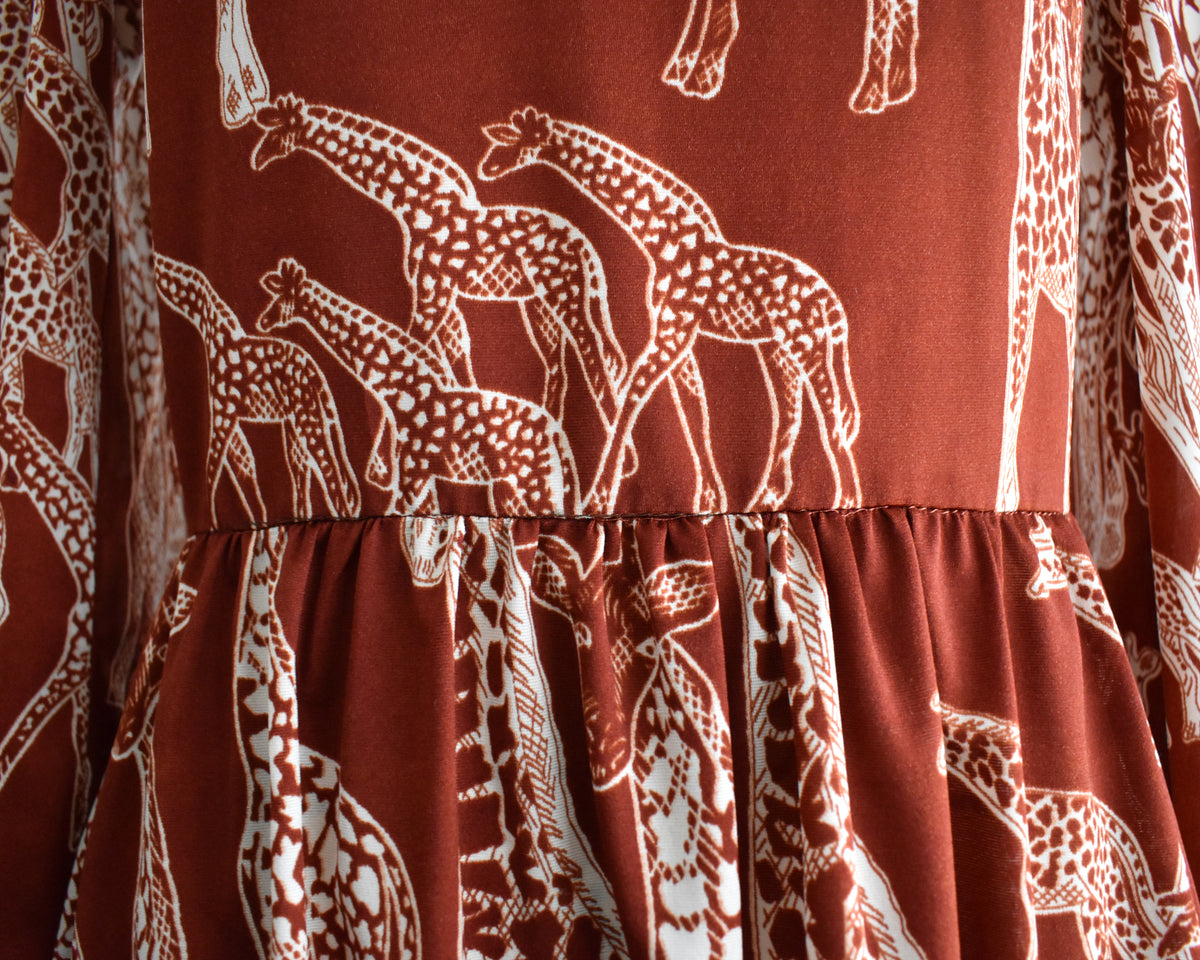 close up of the waist of a vintage 1970s long-sleeve A-line dress displayed on a mannequin against a plain white background. The dress features a rust-brown base with a white giraffe print and a keyhole neckline with button details.