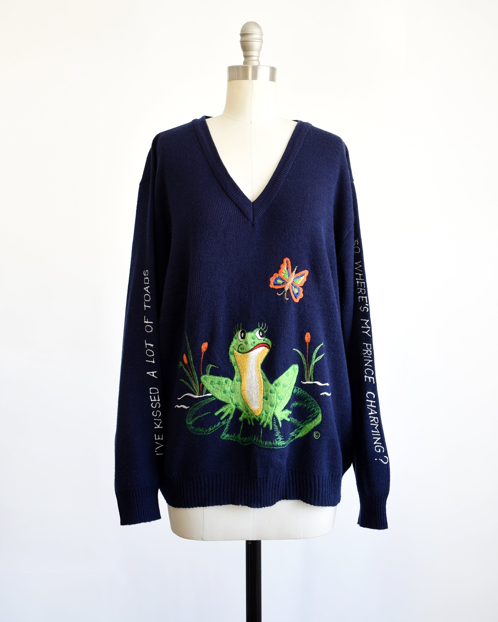 a vintage navy blue 1970s embroidered frog and butterfly sweater that says in white embroidery on the right sleeve "I've Kissed A Lot Of Frogs," and the left sleeve has white embroidery "So Where Is My Prince Charming?"