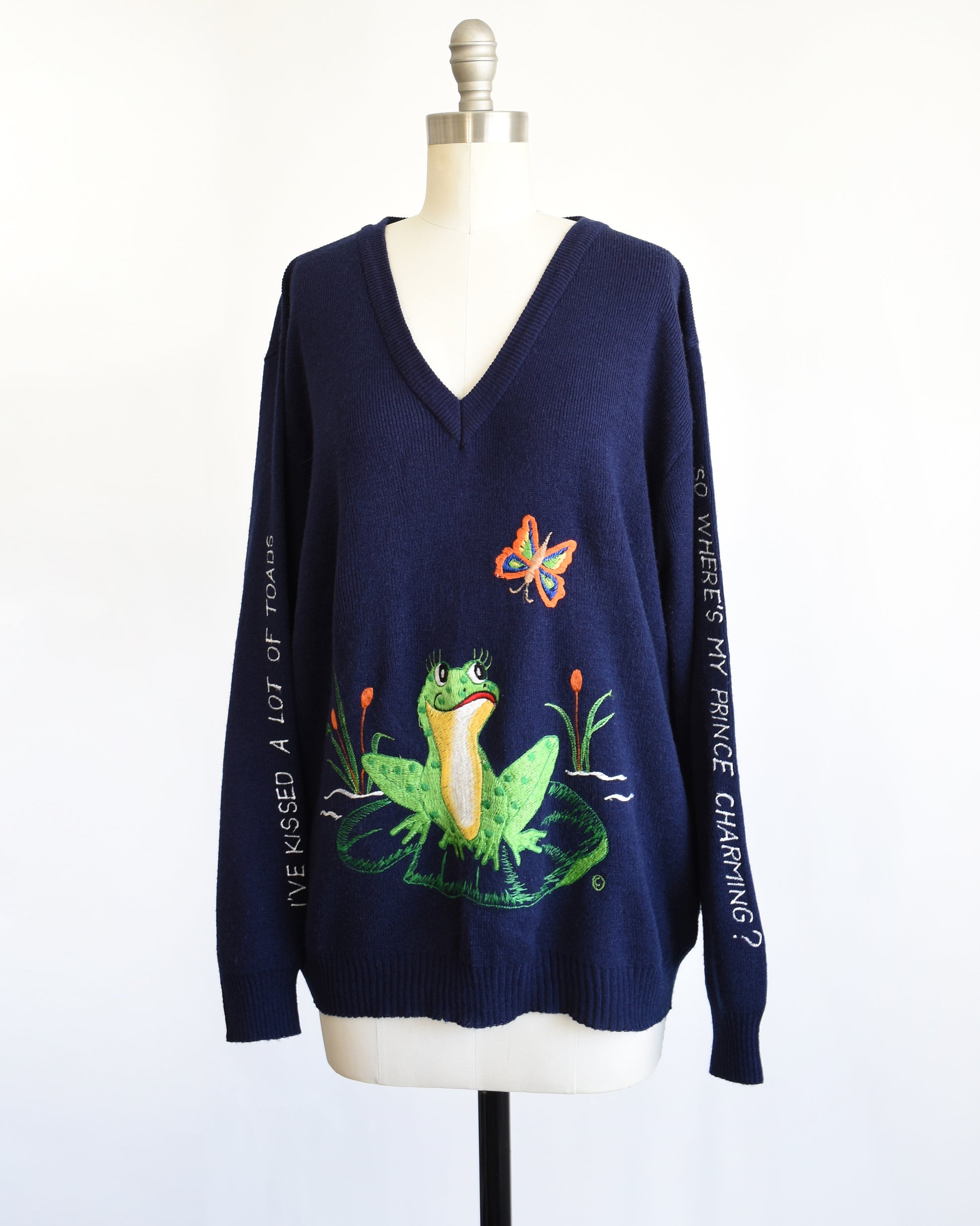 side front view of a vintage navy blue 1970s embroidered frog and butterfly sweater that says in white embroidery on the right sleeve "I've Kissed A Lot Of Frogs," and the left sleeve has white embroidery "So Where Is My Prince Charming?"