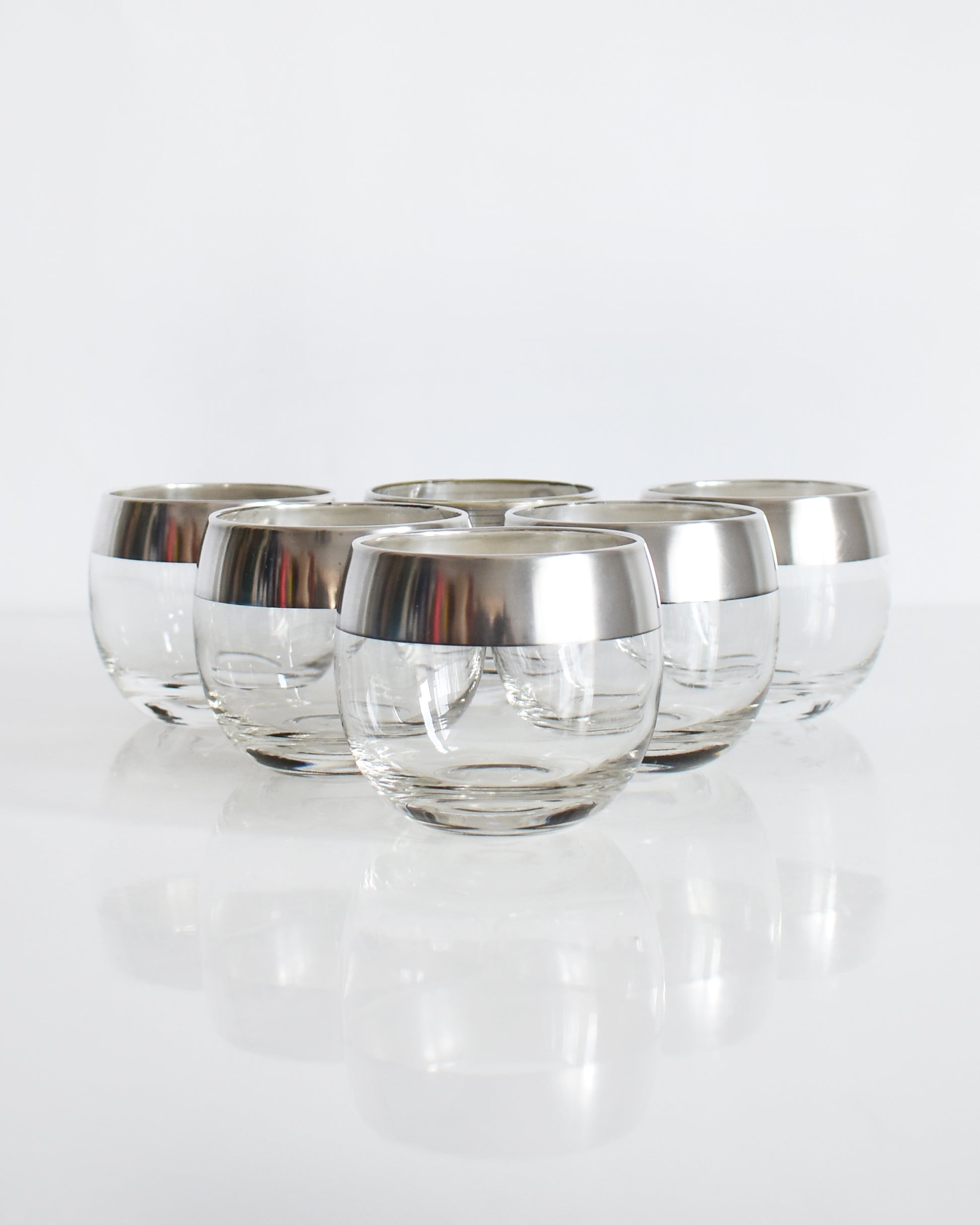 A row of six silver rim roly poly glasses that are round with flat bottoms against a white background