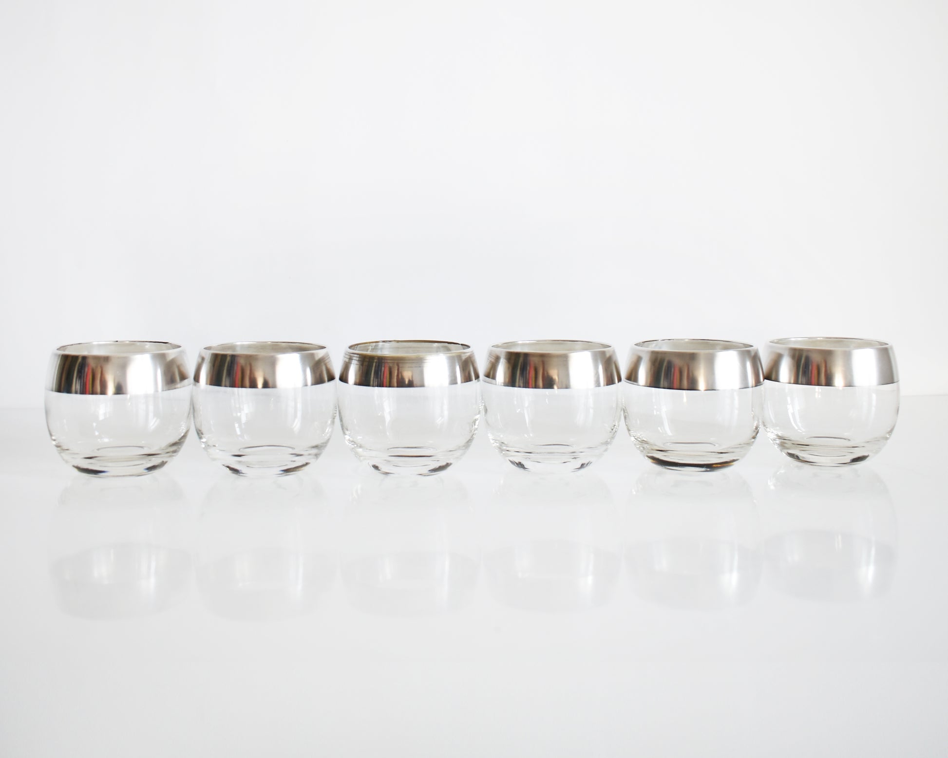 A row of six silver rim roly poly glasses that are round with flat bottoms against a white background