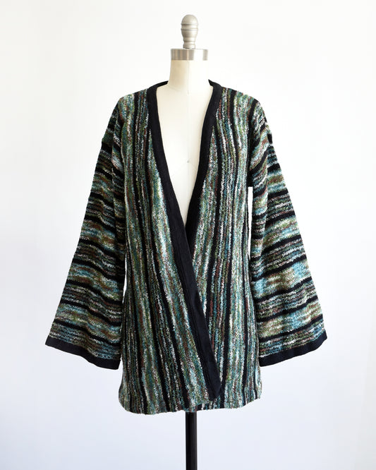 A vintage 1970s open-front cardigan with wide bell sleeves, featuring a textured vertical stripe pattern in black, green, and white, displayed on a white dress form mannequin.