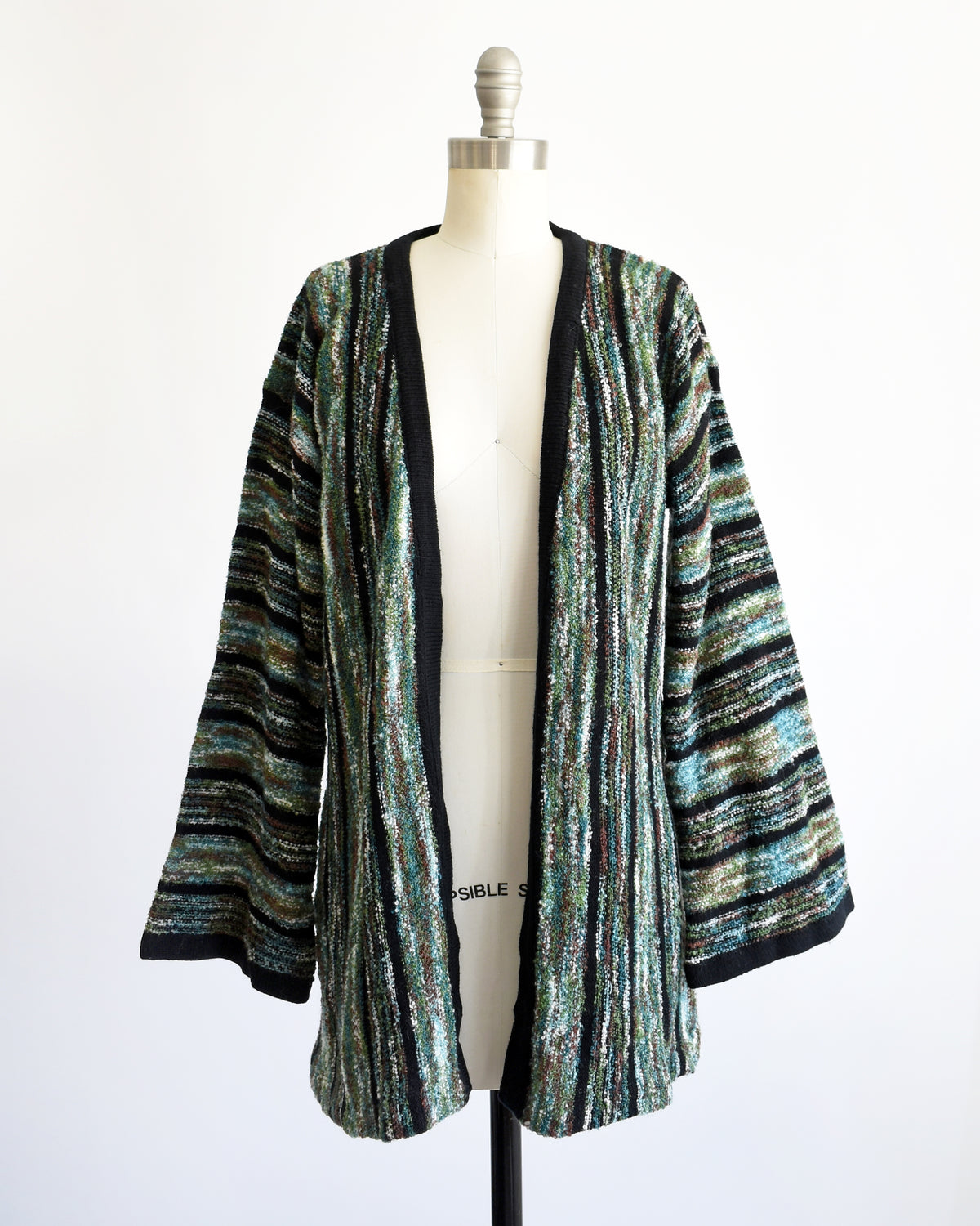 A vintage 1970s open-front cardigan with wide bell sleeves, featuring a textured vertical stripe pattern in black, green, and white, displayed on a white dress form mannequin.