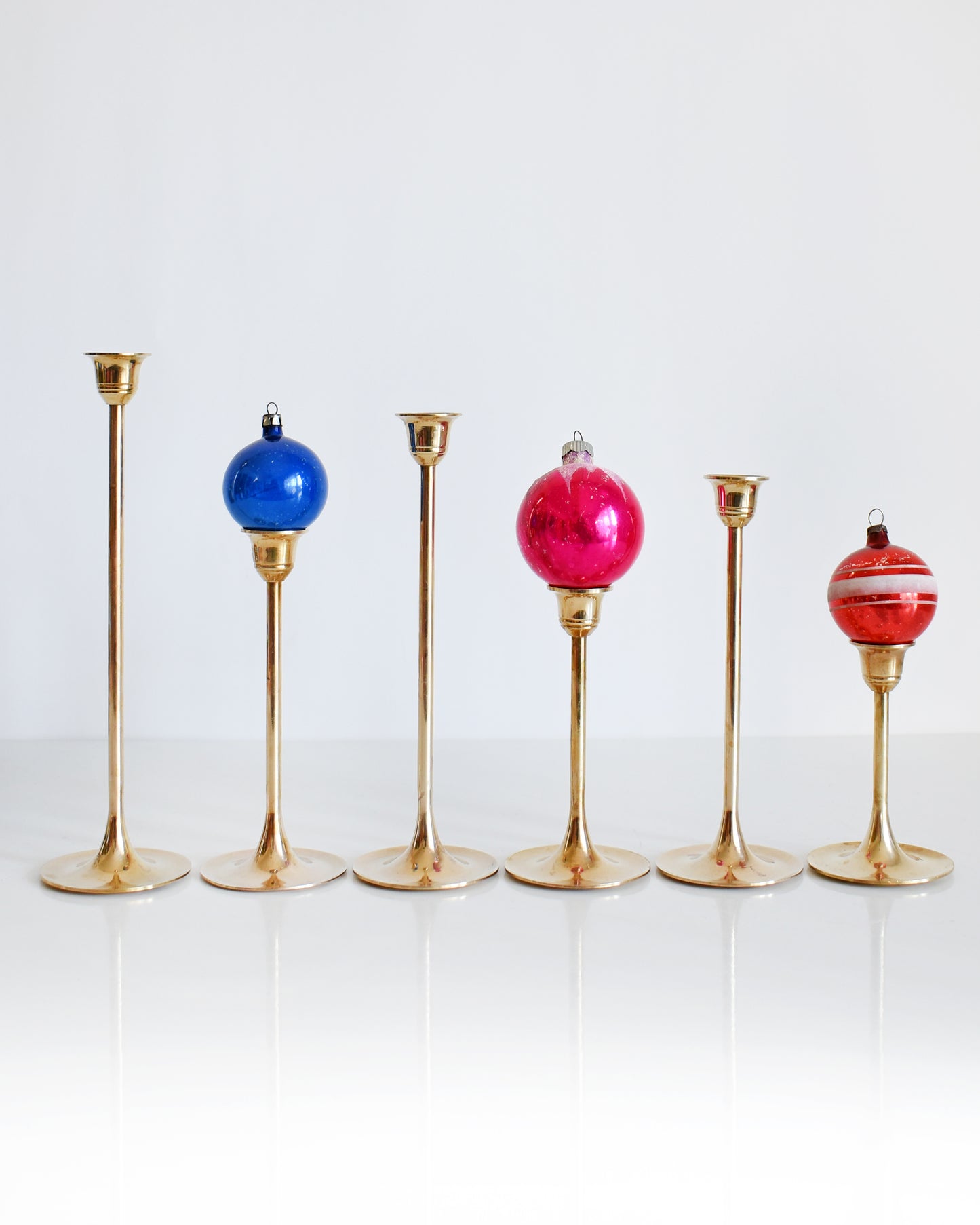 A vintage set of six of different vintage brass graduated candlesticks. Each of the candlesticks have tapered tulip shaped holders, thin stems, and a round base. There are three vintage holiday ornaments on three of the candlesticks.