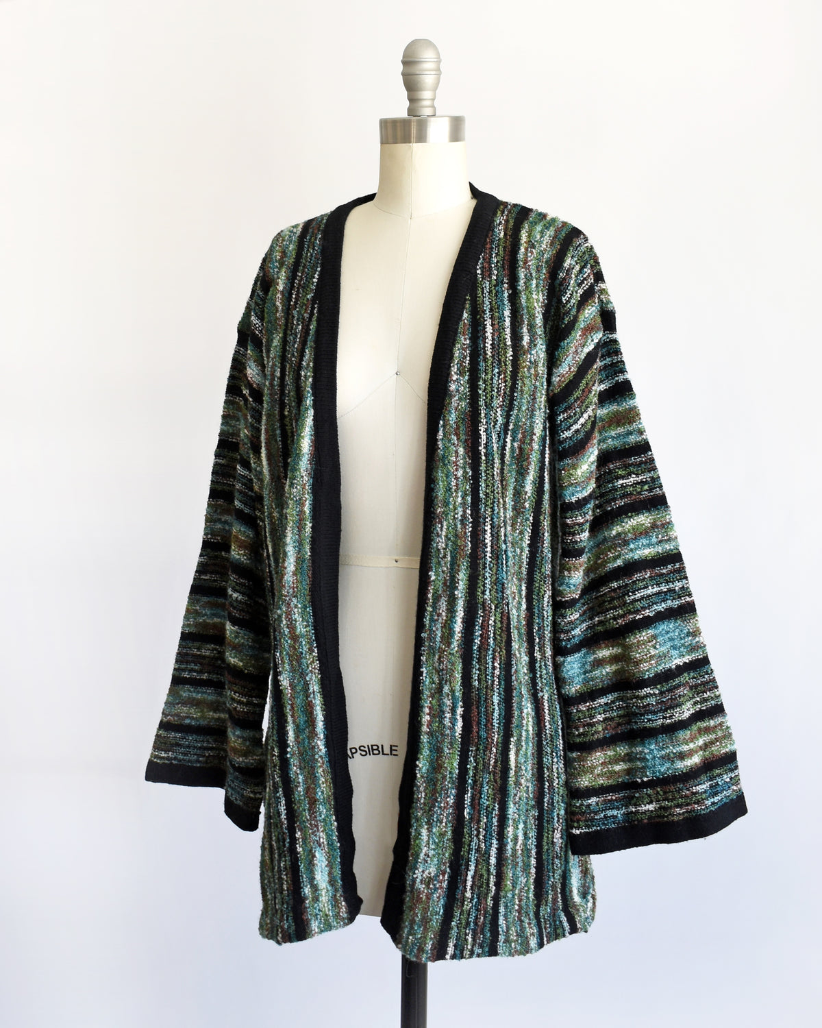 side front view of a vintage 1970s open-front cardigan with wide bell sleeves, featuring a textured vertical stripe pattern in black, green, and white, displayed on a white dress form mannequin.