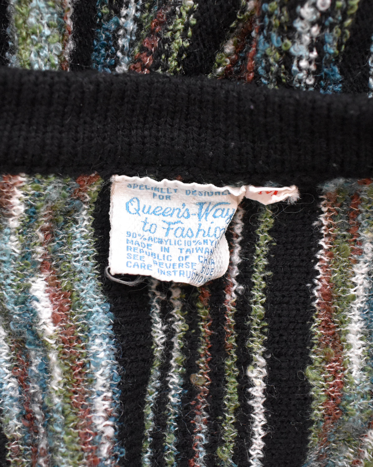 Close-up of a vintage striped knit sweater with a visible sewn-in label reading 'Queen's Way to Fashion' and 'Specialty Designed for.' The sweater has vertical stripes in black, blue, green, white, and rust red, with a thick black ribbed band across the top.