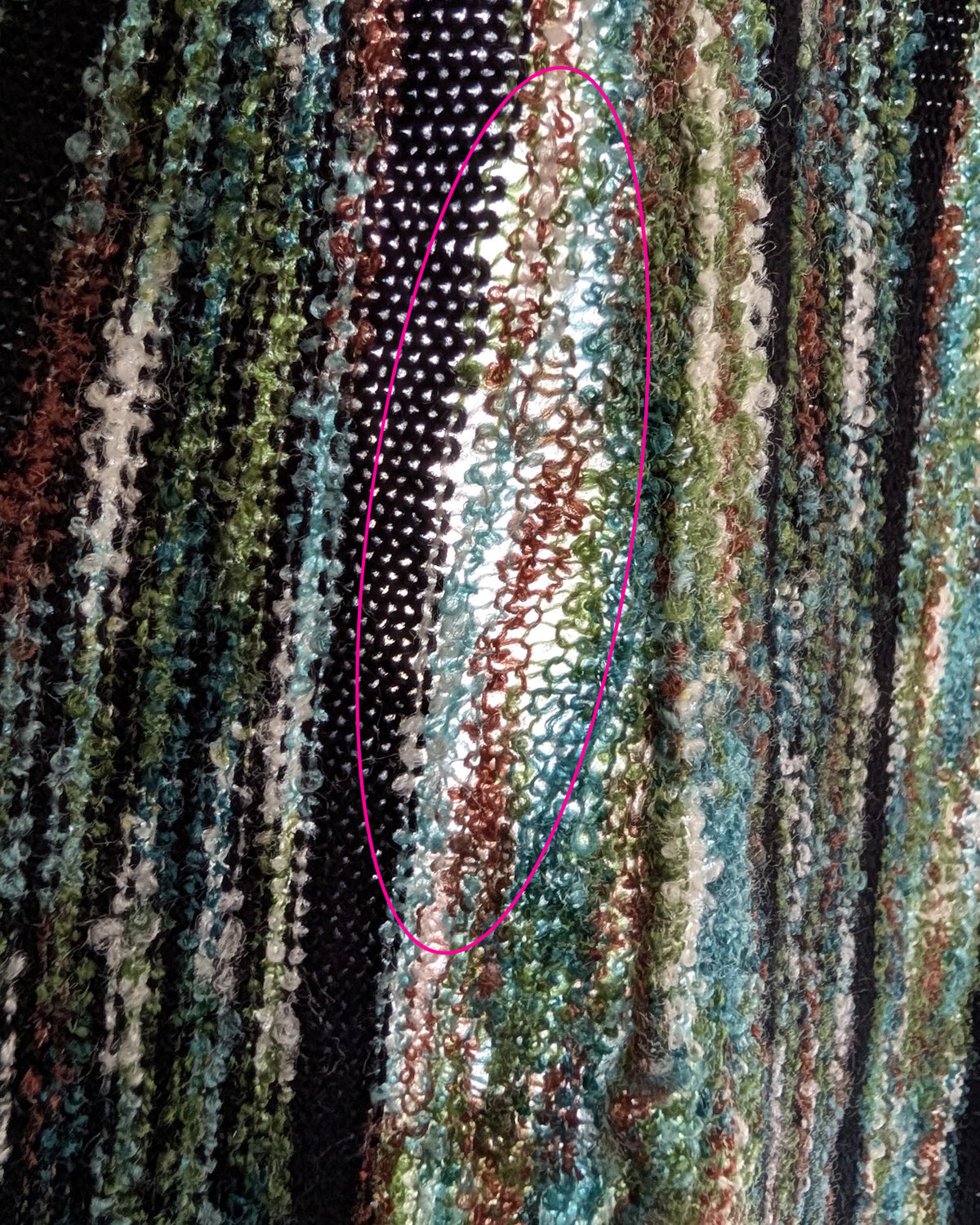 Close-up of a textured, multicolored knit fabric with vertical stripes in black, green, teal, brown, and cream. The highlighted pink oval shows a loose weave.