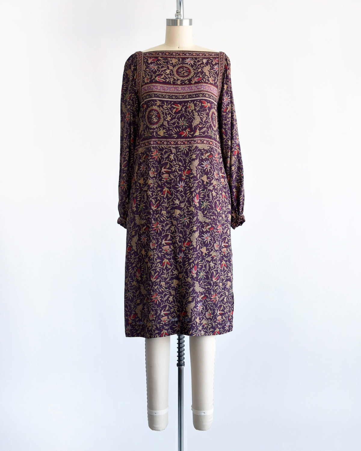 A vintage 1980s long-sleeve knee-length dress with an intricate bohemian-style floral and bird print, displayed on a cream mannequin against a white background.