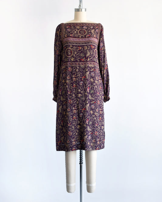 A vintage 1980s long-sleeve knee-length dress with an intricate bohemian-style floral and bird print, displayed on a cream mannequin against a white background.