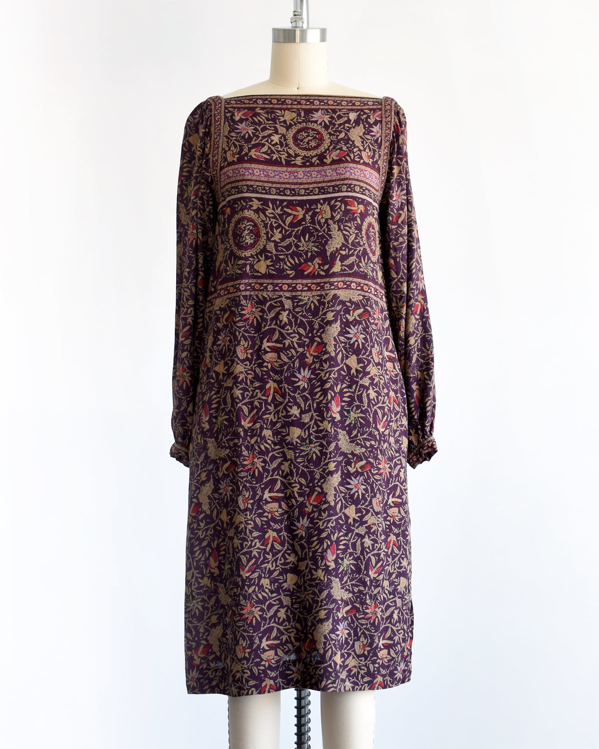A vintage 1980s long-sleeve knee-length dress with an intricate bohemian-style floral and bird print, displayed on a cream mannequin against a white background.