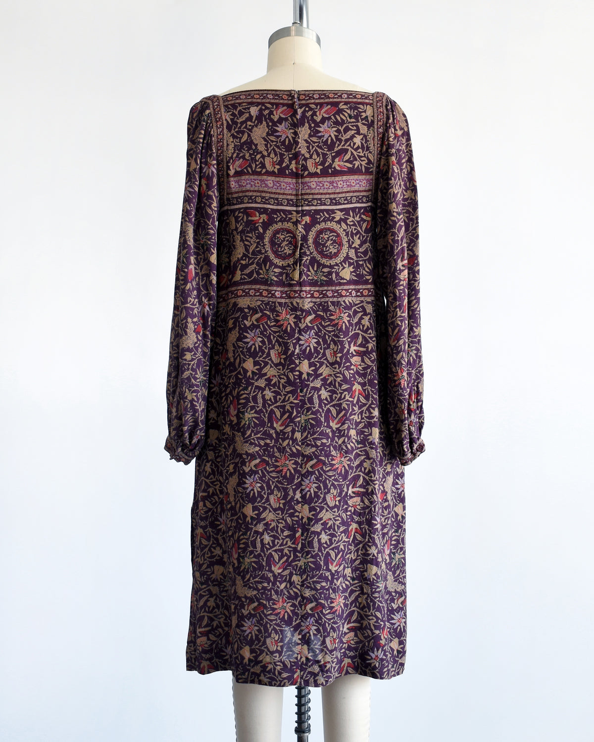back view of a vintage 1980s long-sleeve knee-length dress with an intricate bohemian-style floral and bird print, displayed on a cream mannequin against a white background.