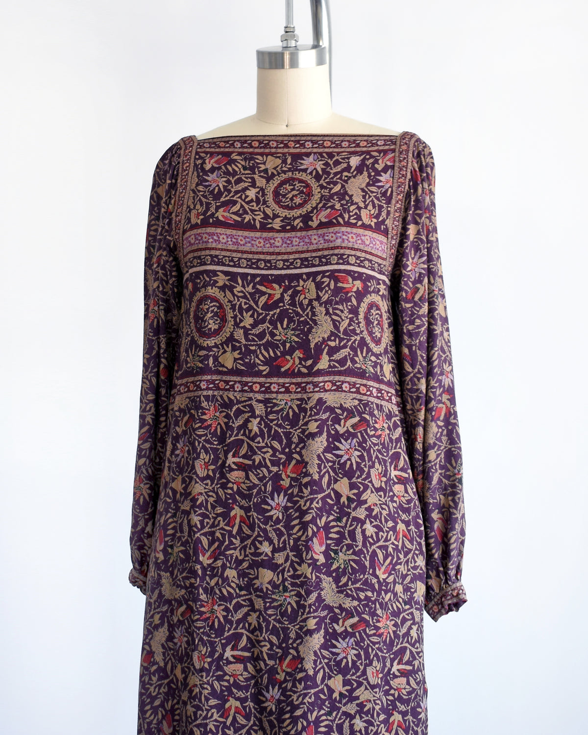 side front view of a vintage 1980s long-sleeve knee-length dress with an intricate bohemian-style floral and bird print, displayed on a cream mannequin against a white background.