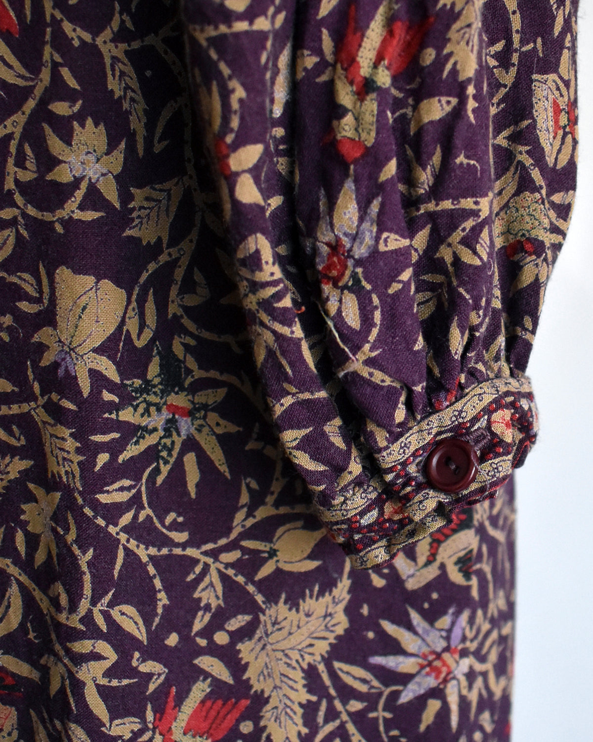 Close-up of a patterned fabric garment, likely a dress or long-sleeve top, featuring a dark purple base with beige botanical motifs and small red floral accents. A matching patterned cuff band with a dark brown button is visible.